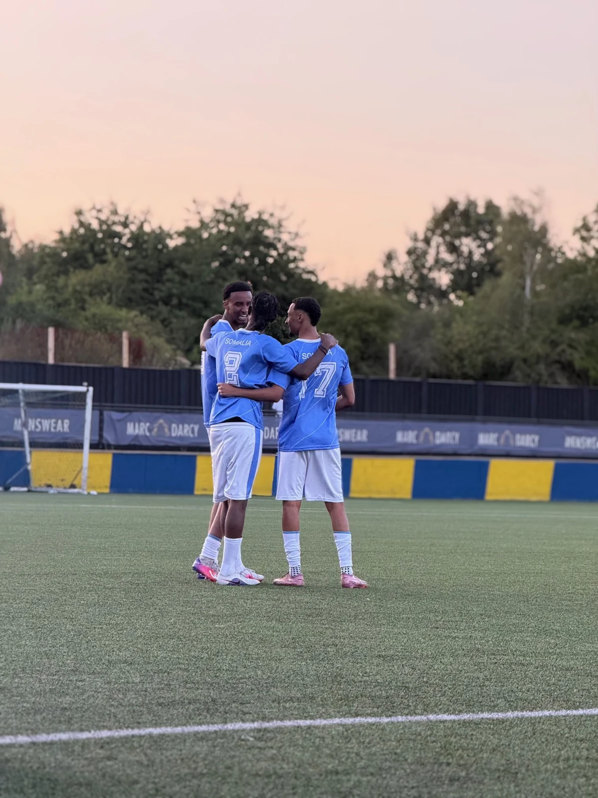 Three Unity Cup players embracing on the pitch at dusk.